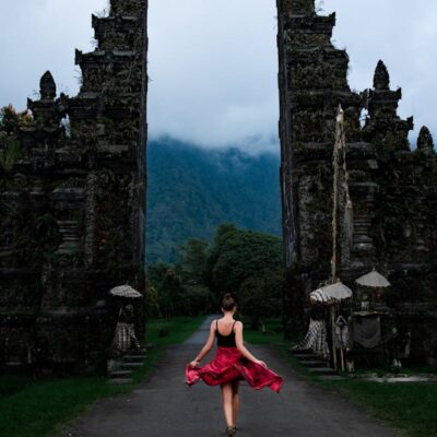 A woman twirls in a red dress at the famous Handara Gate in Bali, Indonesia, amidst misty mountains.